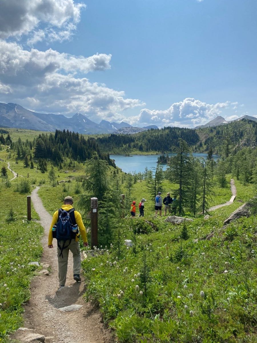 Hikers descending a trail through subalpine meadows toward a turquoise lake