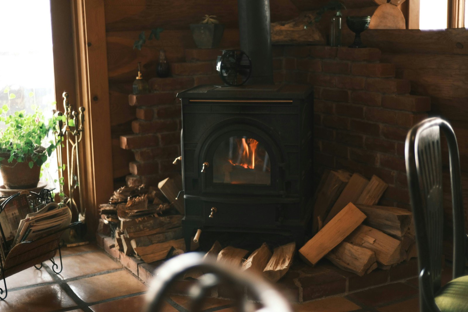 Black woodstove burning beside a stacked woodpile in a log-walled room